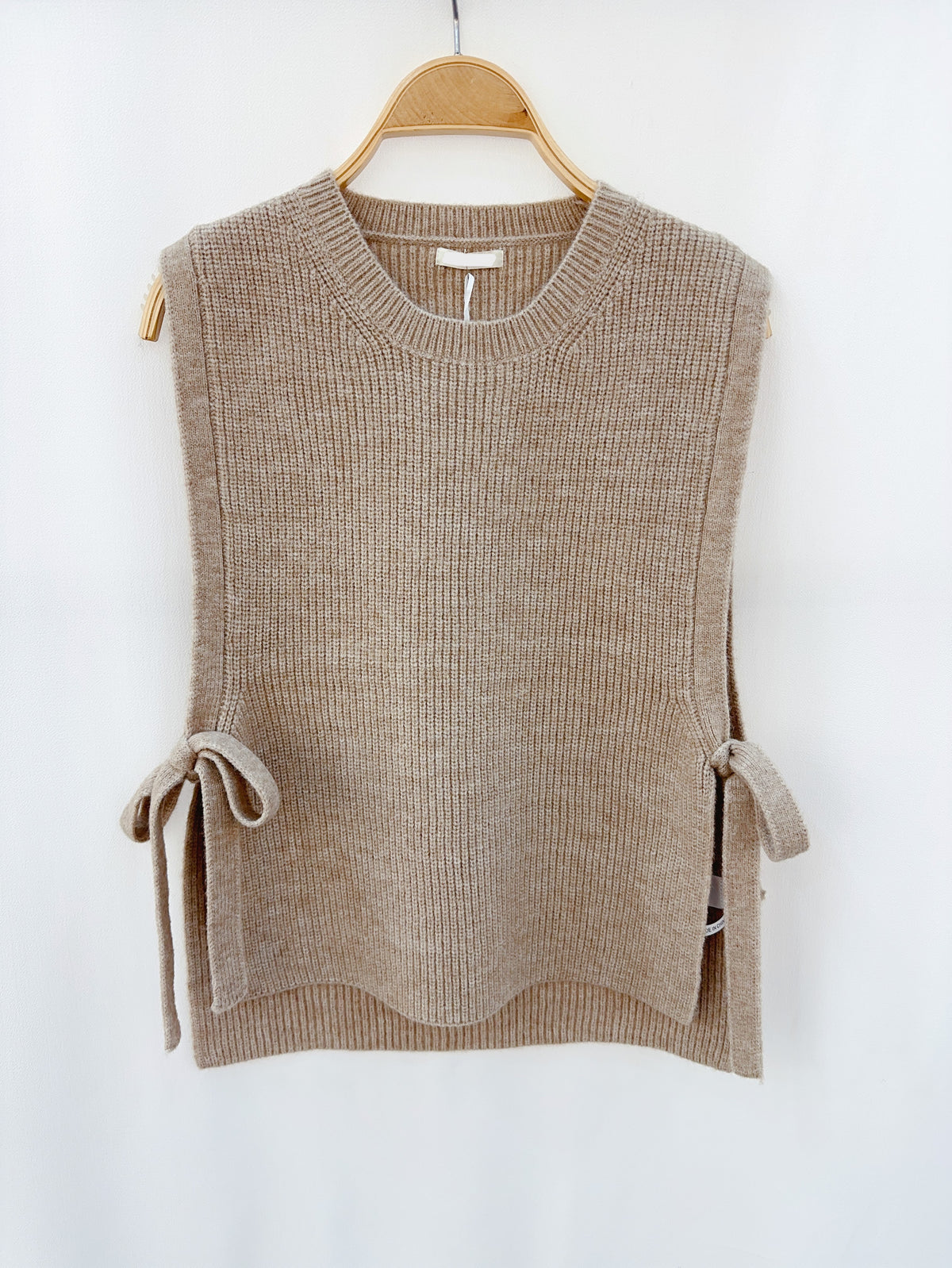 Taupe gebreide mouwloze top met strikdetails, hangend op een houten hanger tegen een neutrale achtergrond.