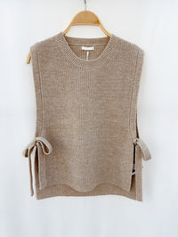 Taupe gebreide mouwloze top met strikdetails, hangend op een houten hanger tegen een neutrale achtergrond.