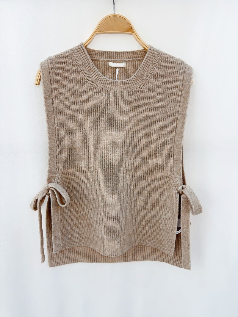 Taupe gebreide mouwloze top met strikdetails, hangend op een houten hanger tegen een neutrale achtergrond.